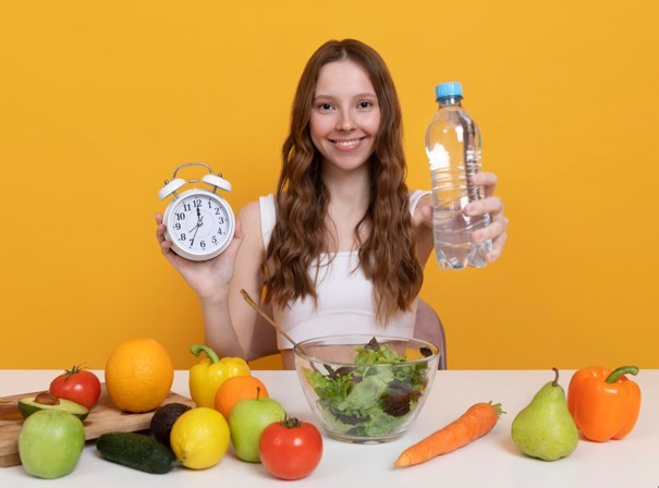 kobieta trzyma wodę i zegarek, przed nią sałata z diety pudełkowej na detoks organizmu oraz owoce i warzywa