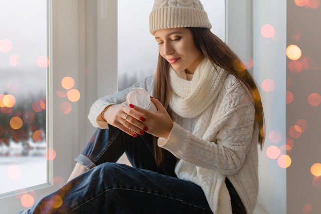 young attractive woman in stylish white knitted sweater, scarf and hat sitting at home on windowsill at Christmas holding glass snow ball present decoration, winter forest view, lights bokeh